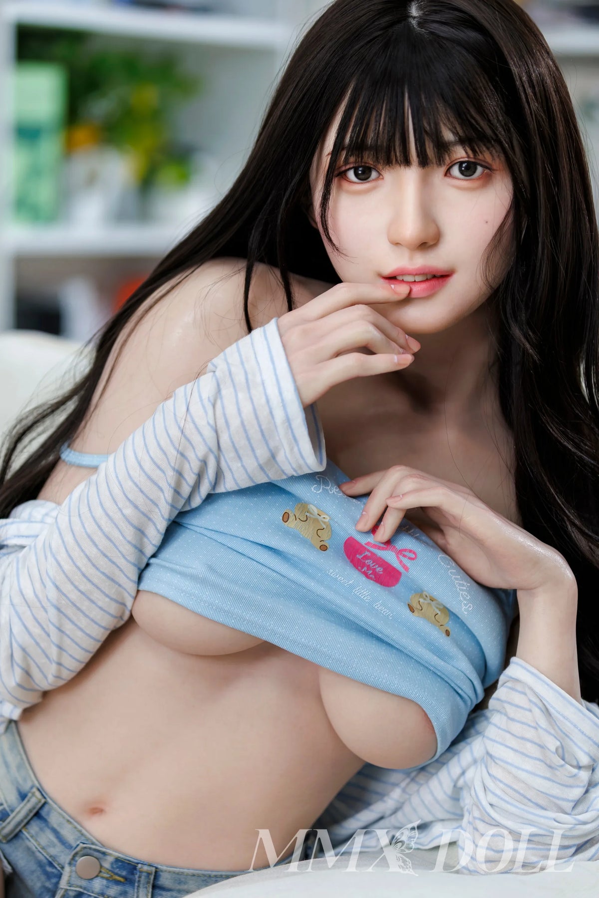 A close-up of a doll or model with long dark hair, wearing a light blue cropped top with cartoon graphics and a white-and-blue striped long-sleeve shirt, posed indoors against a softly blurred background. A watermark reading 'MMX DOLL' appears at the bottom.