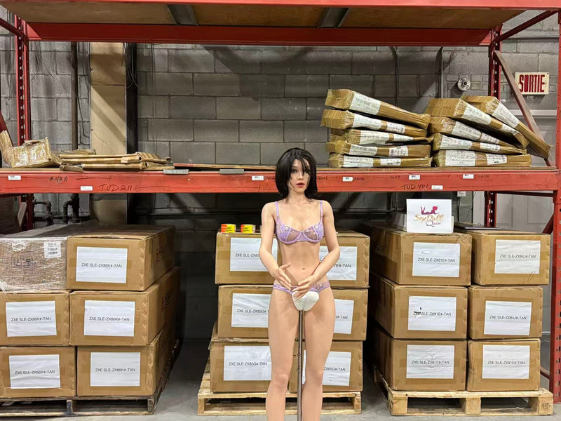 A sex doll wearing a light purple lace bra and white underwear stands in front of a warehouse shelving unit filled with cardboard boxes.