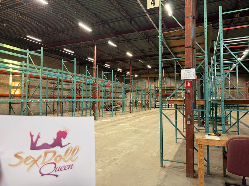 A large warehouse interior with rows of teal and red industrial shelving units on a concrete floor. A white sign in the lower left reads 'Sex Doll Queen' with a purple and gold graphic.