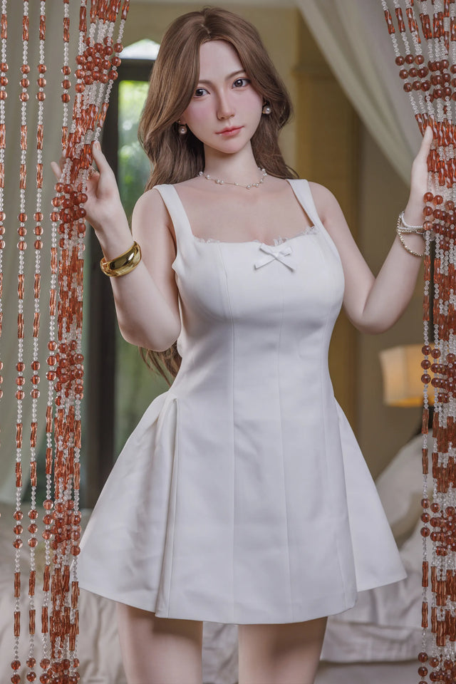 A doll wearing a white sleeveless mini dress with a small bow detail at the neckline, standing in front of a beaded curtain in an indoor setting.